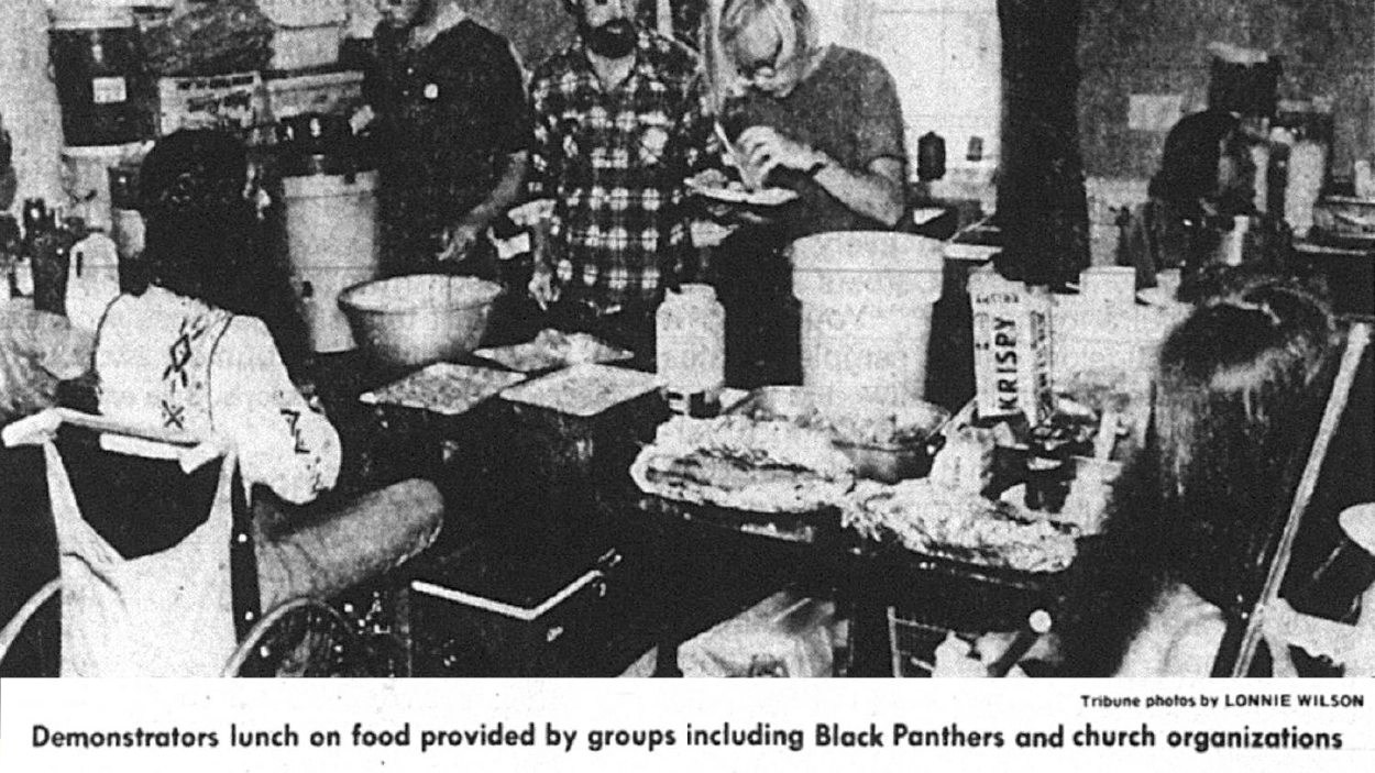 A black and white photograph of people serving, and being served food indoors in a small room. One of the people being served is in a wheelchair. The photo is captioned, 'Demonstraters lunch on food provided by groups including Black Panthers and church organizations.' The photo is attributed to Lonnie Wilson of the Oakland Tribune.