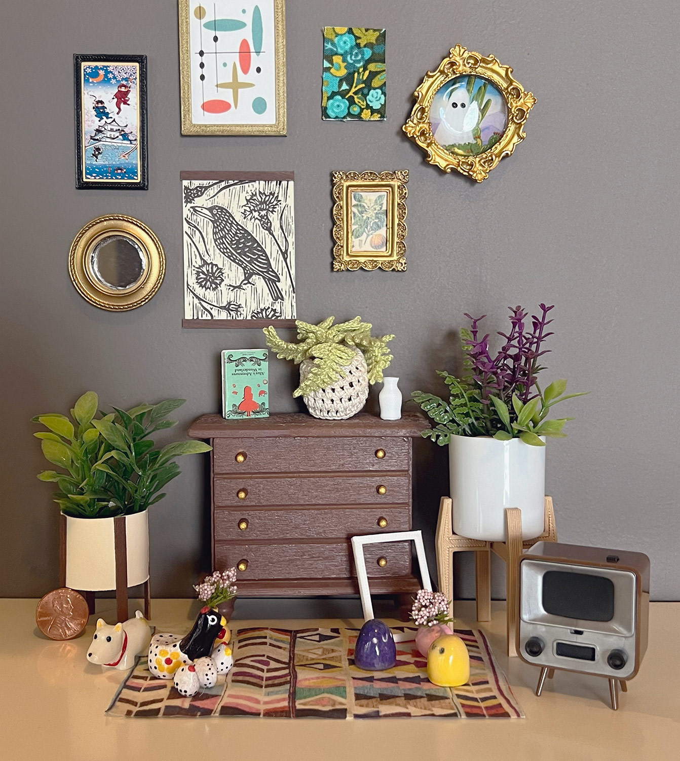 A cozy-looking minature living room placed on the shelf of a piece of normal-sized furniture. The living room has a tiny wooden dresser bureau with gold knobs. A book, vase, and a hand-knit potted fern sit atop the dresser. To the left and right of it are two potted plants, one reminiscent of a monstera and the other a snake plant. In front of the bureai is a small square of fabric decorated with abstract geometric shapes, serving as a carpet. Placed on the carpet are a mini white dog, a mini chicken with three even smaller chicks, two little blobs with smiley faces, and a tiny vase holding angel's breath flowers. There is Above the bureau is a proportinately small decorative mirror and five pieces of art, some with ornate golden frames. In front of the bureau and to the left of the carpet is the tiny TV. It looks like how a TV from the 1940s might, with a large boxy frame, faux wood exterior, and a front metal speaker grill flanked by two control dials. The TV's frame is also held up off the ground via four small faux wooden legs. Finally, a penny is placed on the side of the carpet opposite of the TV, to better communicate the sense of scale.