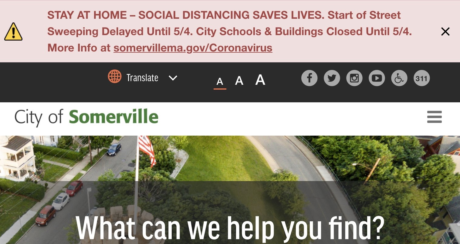 A pink banner taking up one quarter of the screenshot of the City of Somerville homepage. The banner has an alert icon, body copy, and a close button. The body copy reads, 'Stay at home - social distancing saves lives. Start of Street Sweeping Delayed Until 5/4. City Schools & Buildings Closed Until 5/4. More Info at somervillema.gov forward slash Coronavirus'.