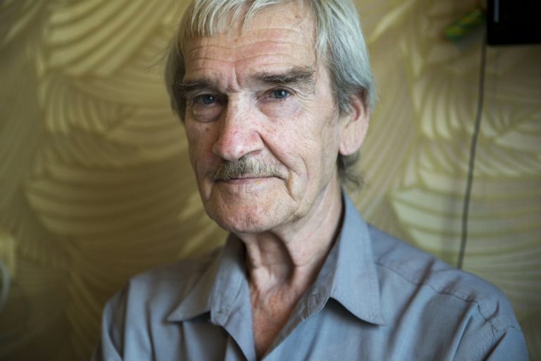 A thin, older white man with salt and pepper gray hair and moustache. He is staring at the camera with a knowing, inscrutable expression.